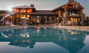 Large modern wooden house with illuminated windows, stone accents, and outdoor pool, viewed from poolside in the evening—an ideal setting to discuss sudden wealth management with your trusted advisor amidst stylish patio furniture.